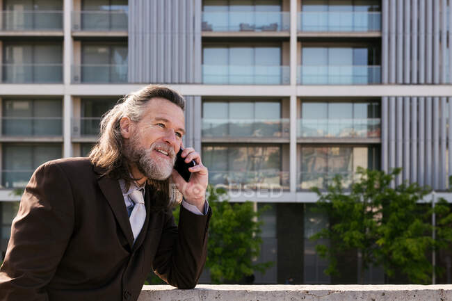 Вид сбоку улыбающийся бородатый взрослый мужчина с седыми волосами в стильном костюме, обсуждающий деловые вопросы во время телефонного разговора, стоя напротив современных городских зданий — стоковое фото