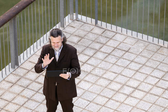 Сверху бородатый мужчина средних лет в элегантном костюме машет рукой, приветствуя партнера во время видеовстречи через цифровой планшет на террасе — стоковое фото