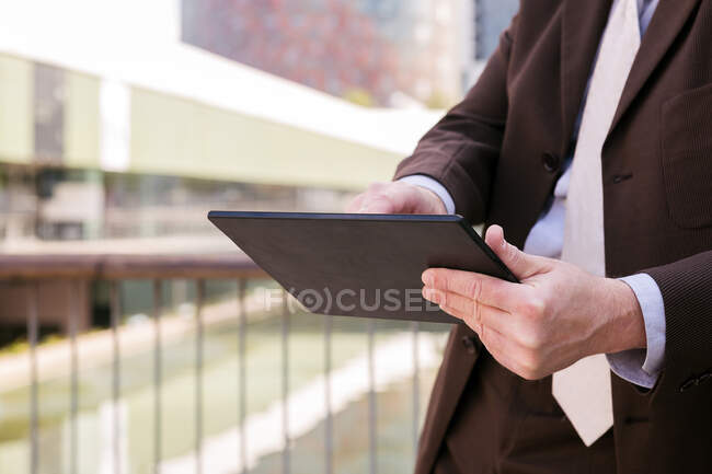 Cropped unrecognizable man in formal outfit browsing tablet while standing on embankment in city — Stock Photo