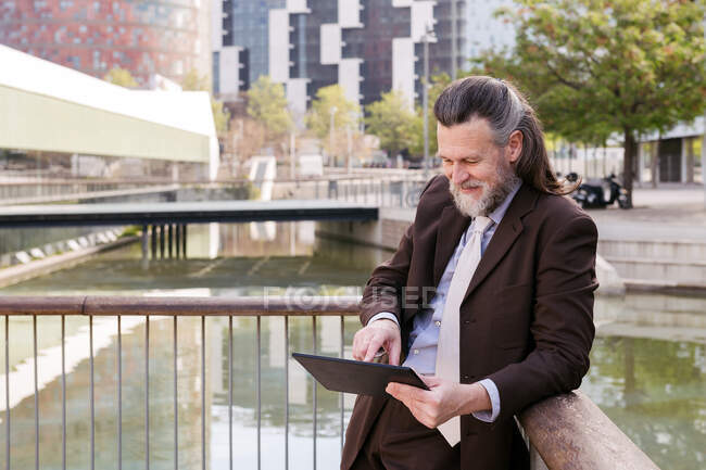 Позитивний респектабельний бородатий чоловік середнього віку в офіційному вбранні планшета, стоячи на набережній в місті — стокове фото