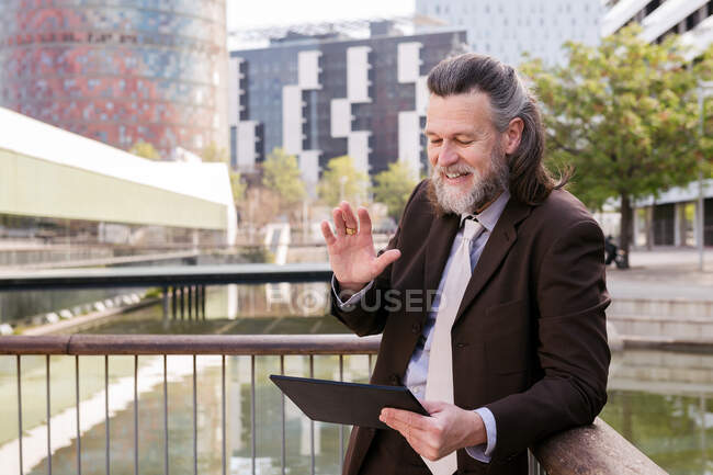 Glücklicher bärtiger Mann mittleren Alters im eleganten Anzug winkt mit der Hand, während er Partner beim Videotreffen via digitalem Tablet auf der Terrasse begrüßt — Stockfoto