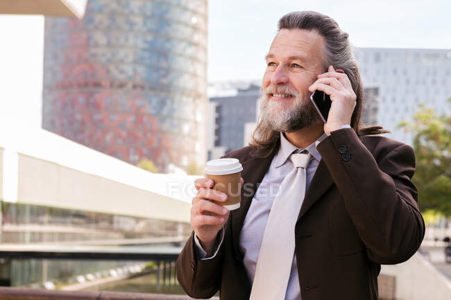 Sonriente hombre barbudo maduro en elegante traje elegante con taza de café para llevar en la mano hablando por teléfono móvil mientras está de pie en la calle urbana - foto de stock