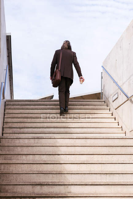 From below back view of unrecognizable stylish businessman in classy suit with briefcase and takeaway coffee climbing up stairs in city — Stock Photo