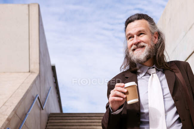 From below of confident mature male executive manager in classy suit with bag drinking takeaway coffee while walking down stairs in city — Stock Photo