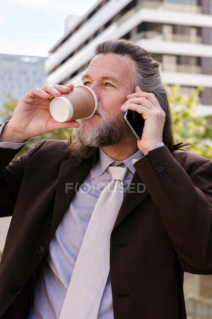 Sonriente hombre barbudo maduro en elegante traje elegante con taza de café para llevar en la mano hablando por teléfono móvil mientras está de pie en la calle urbana - foto de stock