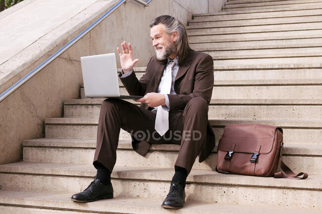 Homem barbudo envelhecido positivo em terno elegante situado na escada e acenando a mão e dizendo olá ao ter reunião de vídeo on-line via laptop — Fotografia de Stock