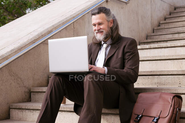 Empresario masculino barbudo de mediana edad positivo en ropa formal sentado en la escalera y trabajando en línea en el ordenador portátil en la ciudad - foto de stock