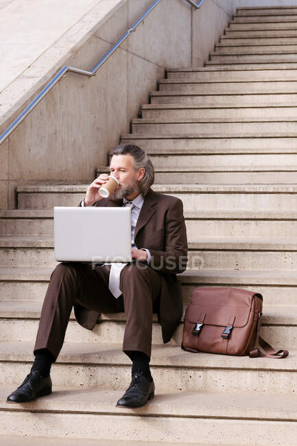 Empresario masculino barbudo de mediana edad en ropa formal sentado en la escalera bebiendo café para llevar y trabajando en línea en la computadora portátil en la ciudad - foto de stock