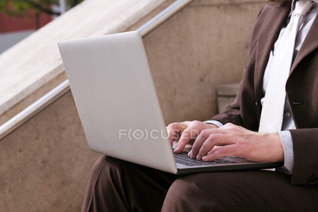 Cropped unrecognizable male entrepreneur in formal clothes sitting on stairway and working online on laptop in city — Stock Photo
