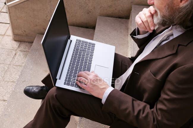 Обрезанный неузнаваемый мужчина-предприниматель в формальной одежде сидит на лестнице и работает онлайн на ноутбуке в городе — стоковое фото