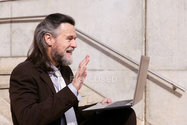 Вид збоку позитивного старого бородатого чоловіка в елегантному костюмі, що сидить на сходах і махає рукою і привітається, маючи онлайн-відео-зустріч через ноутбук — стокове фото