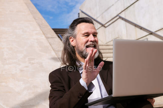 From below positive aged bearded male in elegant suit siting on stairway and waving hand and saying hello while having online video meeting via laptop — Stock Photo