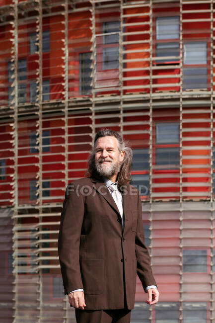 Hombre barbudo de mediana edad positivo en traje de estilo empresarial mirando hacia otro lado pensativamente contra el edificio urbano - foto de stock