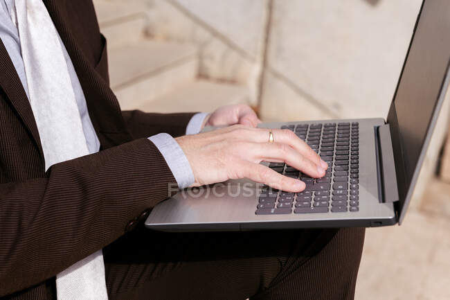 Cortado empresario masculino irreconocible en ropa formal sentado en la escalera y trabajando en línea en el ordenador portátil en la ciudad - foto de stock