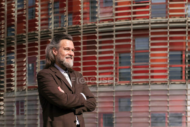 Side view of positive middle aged bearded man in business style outfit standing with arms crossed and looking away pensively against urban building — Stock Photo