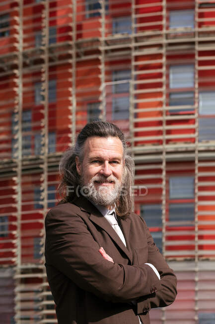Side view of positive middle aged bearded man in business style outfit standing with arms crossed and looking at camera pensively against urban building — Stock Photo