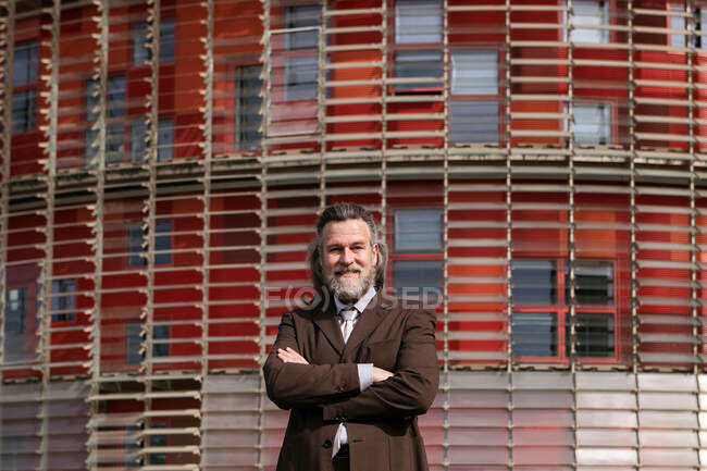 Positive middle aged bearded man in business style outfit standing with arms crossed looking at camera pensively against urban building — Stock Photo
