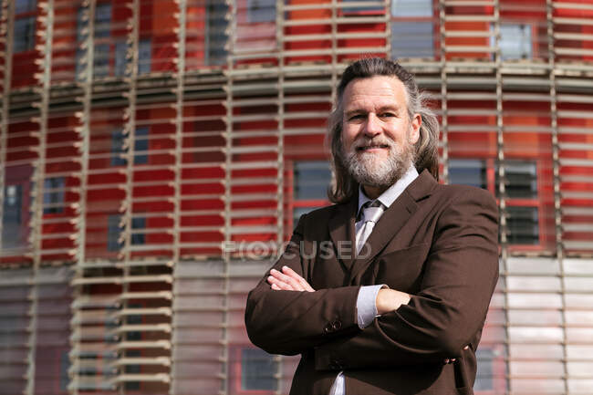 Positive middle aged bearded man in business style outfit standing with arms crossed looking at camera pensively against urban building — Stock Photo