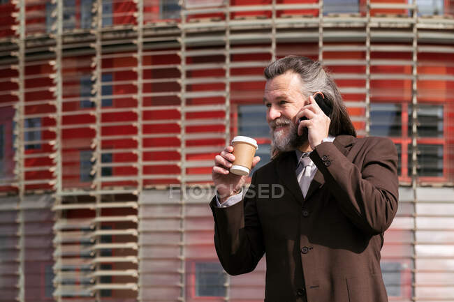Gray haired bearded man in formal suit with cup of takeaway coffee in hand speaking on mobile phone while standing on urban street — Stock Photo