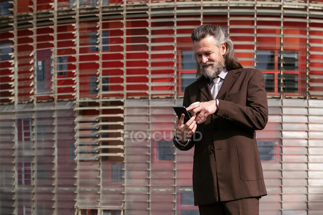 Homme barbu aux cheveux gris en costume formel buvant du café à emporter et naviguant sur son téléphone portable dans la rue urbaine — Photo de stock
