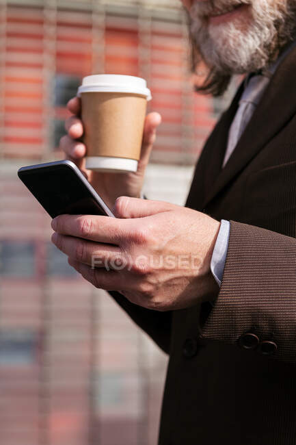 Vista lateral de la cosecha irreconocible hombre barbudo de pelo gris en traje formal beber café para llevar y navegar por el teléfono móvil en la calle urbana - foto de stock
