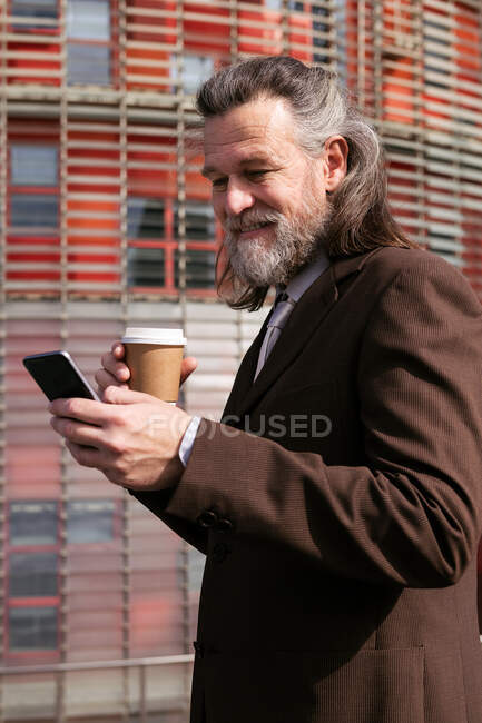Vista lateral do homem barbudo de cabelos grisalhos em terno formal bebendo café takeaway e navegando telefone celular na rua urbana — Fotografia de Stock