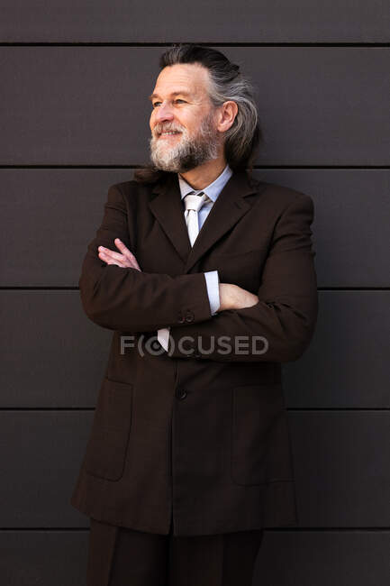 Confiante de sucesso maduro barbudo macho em terno elegante de pé com os braços cruzados e olhando para longe com sorriso contra a parede cinza — Fotografia de Stock