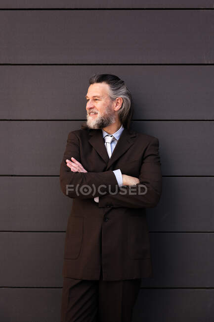 Confident successful mature bearded male in elegant suit standing with arms crossed and looking away with smile against gray wall — Stock Photo