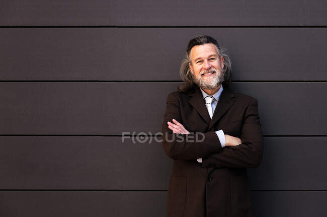 Confiante de sucesso maduro barbudo macho em terno elegante de pé com os braços cruzados e olhando para a câmera com sorriso contra a parede cinza — Fotografia de Stock