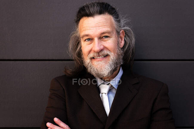 Hombre barbudo maduro con éxito seguro en traje elegante de pie con los brazos cruzados y mirando a la cámara con sonrisa contra la pared gris - foto de stock