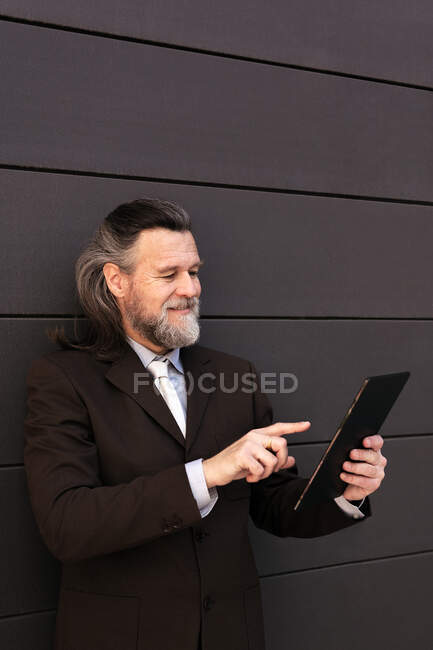 Eleganter bärtiger älterer Mann im eleganten formalen Anzug steht neben grauer Wand und surft auf einem digitalen Tablet im Internet — Stockfoto