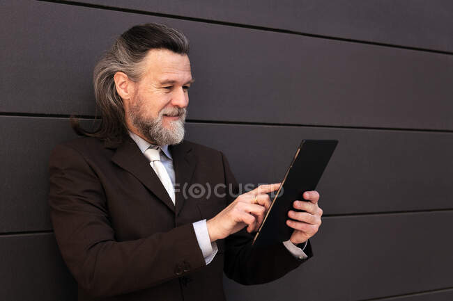 Elegant bearded mature man in classy formal suit standing near gray wall and browsing internet on digital tablet — Stock Photo