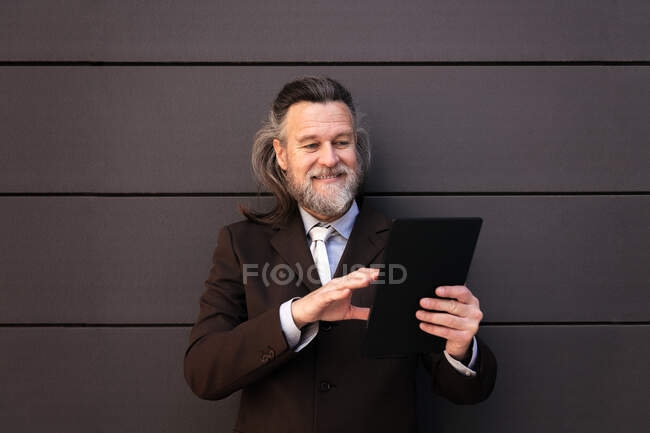 Elegante barbudo hombre maduro en traje formal con clase de pie cerca de la pared gris y navegar por Internet en la tableta digital - foto de stock
