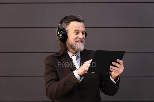Вміст сірого волосся бородатого чоловіка в офіційному костюмі та бездротових навушниках, використовуючи планшет, спілкуючись онлайн проти сірої стіни — стокове фото