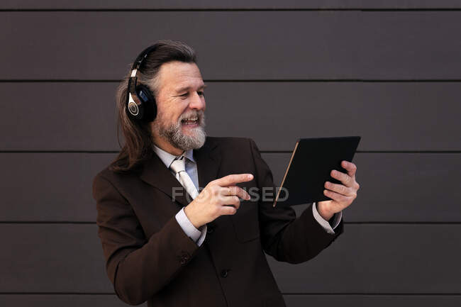 Inhalt grauhaarige bärtige Männchen in formalen Anzug und drahtlose Kopfhörer mit Tablet während der Online-Kommunikation gegen graue Wand — Stockfoto
