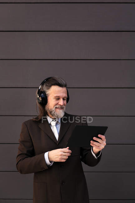 Contenu mâle barbu cheveux gris en costume formel et écouteurs sans fil en utilisant la tablette tout en communiquant en ligne contre le mur gris — Photo de stock