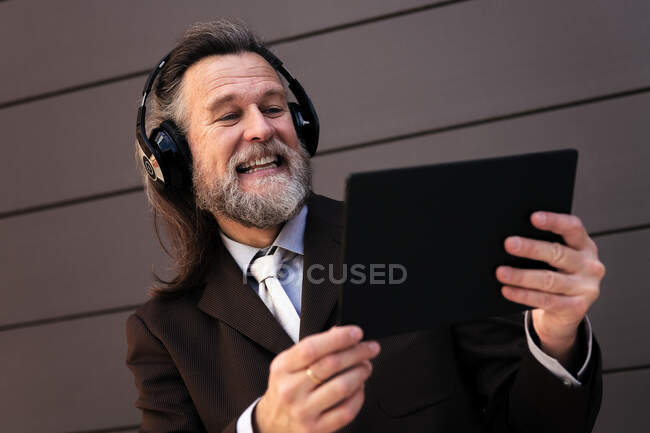 From below content gray haired bearded male in formal suit and wireless headphones using tablet while communicating online against gray wall — Stock Photo