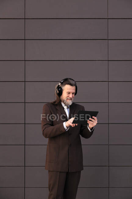 Contenu mâle barbu cheveux gris en costume formel et écouteurs sans fil en utilisant la tablette tout en communiquant en ligne contre le mur gris — Photo de stock