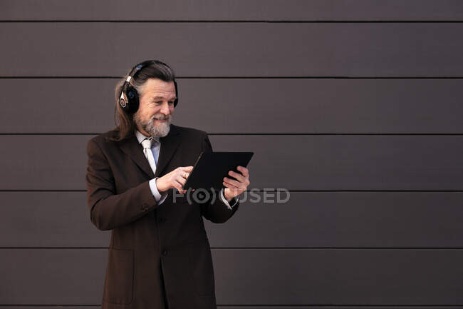 Conteúdo macho barbudo de cabelos grisalhos em terno formal e fones de ouvido sem fio usando tablet enquanto se comunica online contra a parede cinza — Fotografia de Stock