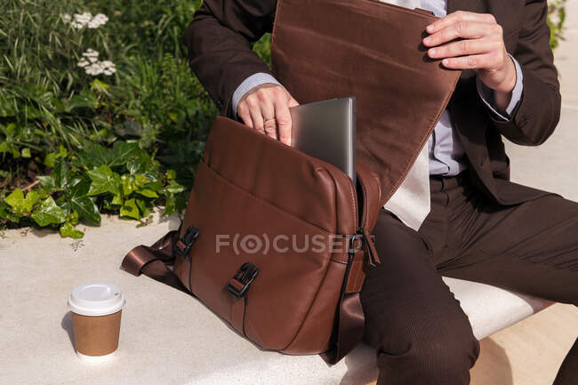 Обрізаний невпізнаваний чоловік-адвокат у формальному одязі, що бере ноутбук з портфеля, сидячи з чашкою кави на міській площі — стокове фото