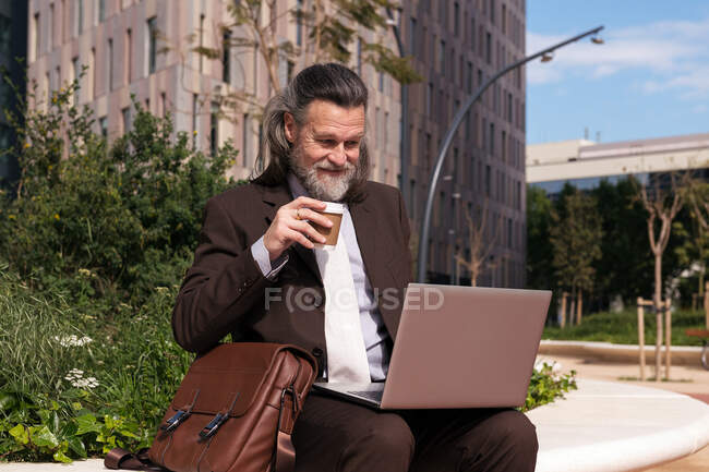 Happy successful gray haired bearded male in elegant suit drinking takeaway coffee using laptop while sitting on urban street — Stock Photo