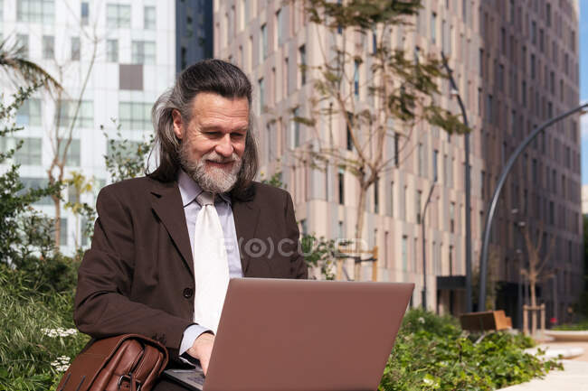 Felice maschio barbuto dai capelli grigi di successo in abito elegante utilizzando il computer portatile mentre seduto sulla strada urbana — Foto stock