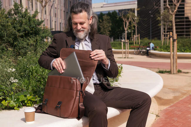 Conteúdo respeitável meia idade barbudo advogado masculino em roupas formais levando laptop de pasta enquanto sentado com xícara de café takeaway na praça urbana — Fotografia de Stock