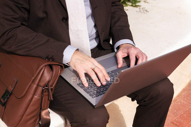 Обрізаний невпізнаваний чоловік в елегантному костюмі, що друкується на ноутбуці, сидячи на міській вулиці — стокове фото