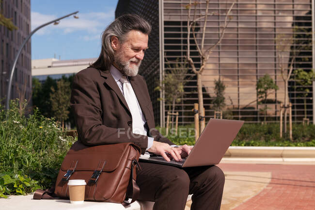 Vista lateral de feliz éxito de pelo gris barbudo macho en traje elegante utilizando el ordenador portátil mientras está sentado en la calle urbana - foto de stock