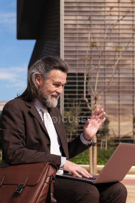 Side view of happy successful gray haired bearded male in elegant suit waving hand and saying hello while having online video meeting via laptop sitting on urban street — Stock Photo