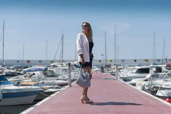 Щаслива жінка в літньому вбранні, що йде вздовж набережної біля моря з швартованими яхтами — стокове фото