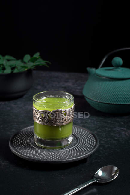 Натюрморт композиція з традиційним східним чаєм мача, подається в скляній чашці з металевим декоративним декором на столі з керамічними чашами та свіжим зеленим листям на чорному тлі — стокове фото