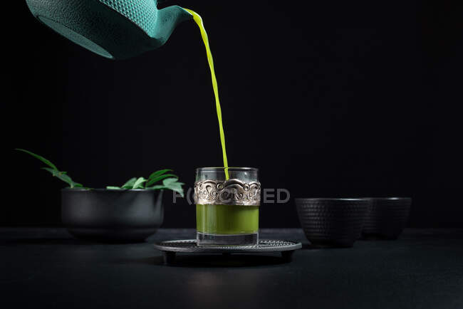 Healthy Japanese matcha tea being poured from green teapot into glass cup with metal ornamental decor during tea ceremony against black background — Stock Photo