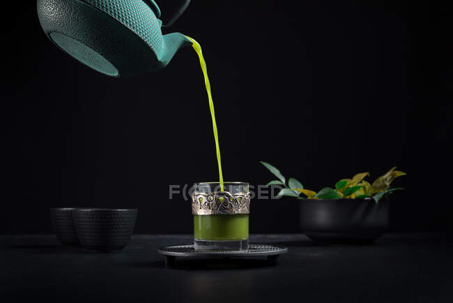 Té matcha japonés saludable que se vierte de la tetera verde en la taza de vidrio con decoración ornamental de metal durante la ceremonia del té contra el fondo negro - foto de stock